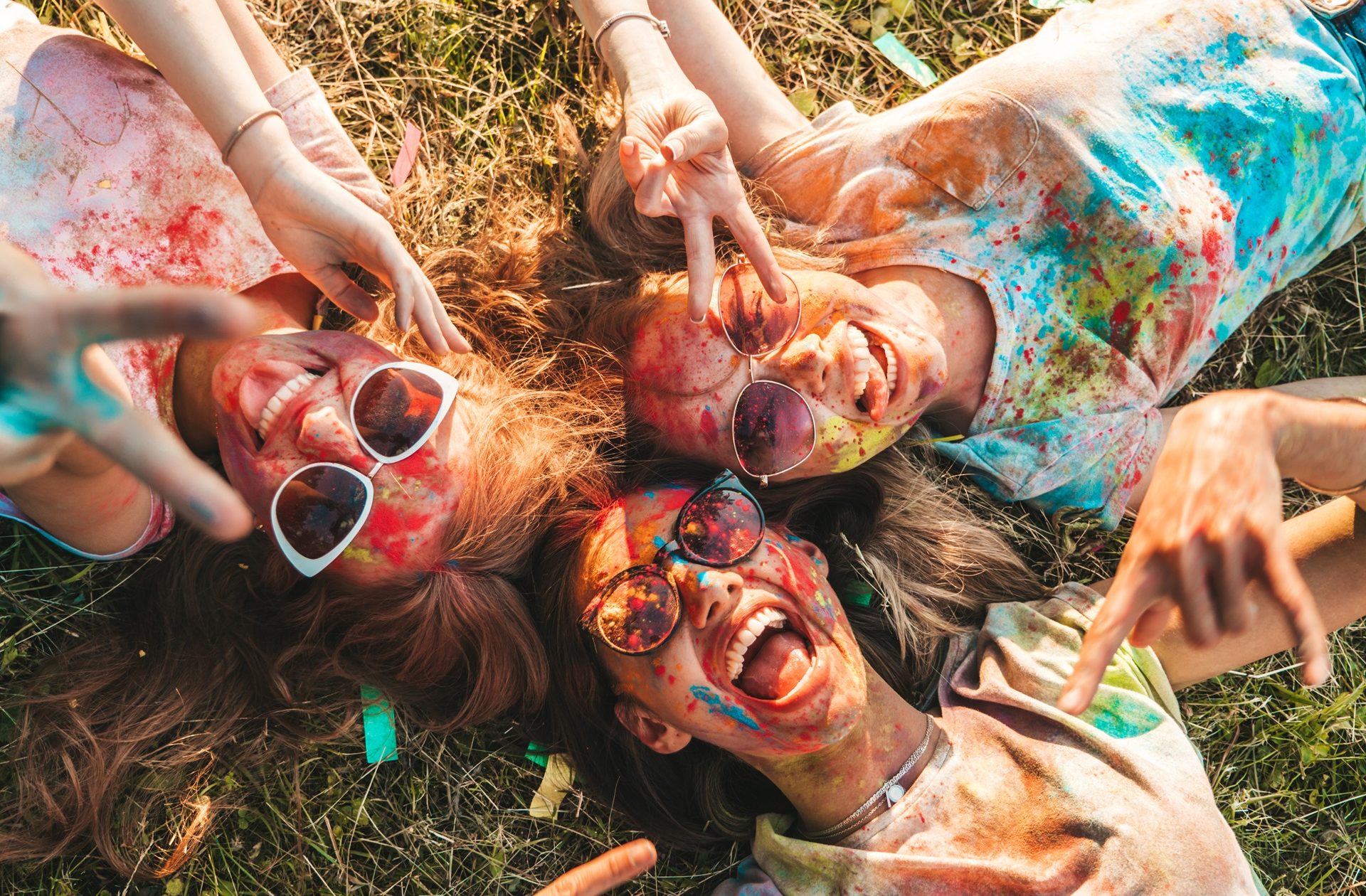 Three happy beautiful girls making party at Holi colors festival in summer time.Young  smiling women friends having fun after music event at sunset.Models lying on the grass in sunglasses